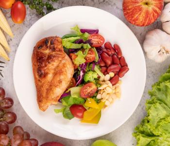 Plate full of healthy food items, including chicken, tomotatoes, lettuce, peppers, and radish. It is surrounded by corn, forks, grapes, garlic, and lettuce on the surface holding the plate